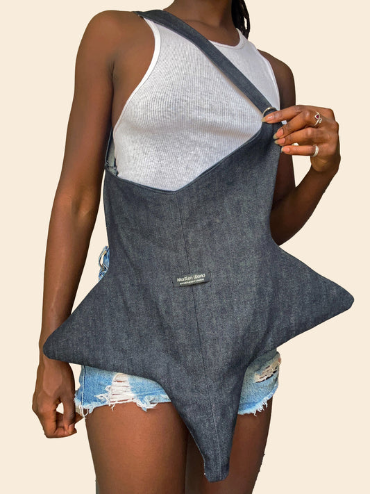 A model wearing a dark gray denim-style star-shaped apron or bib over a white ribbed tank top and distressed denim shorts, holding the top corner of the garment against a plain beige background.